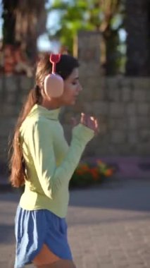 Dışarıda koşan genç bir kadın sarı kapüşonlu, mavi şortlu ve gün ışığında sıcak pembe kulaklıklı..