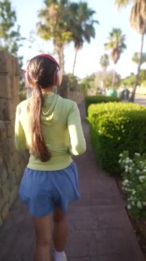 Bir kadın, çevresi bitki ve palmiye ağaçlarıyla çevrili bir yolda kulaklıkla koşarken görülüyor.