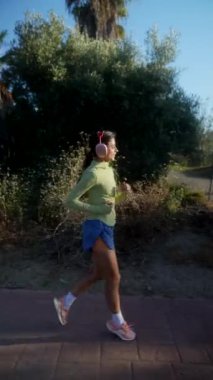 Yeşil bluz ve mavi şort giyen, kulaklık takan bir kadın dışarıda doğal bir ortamda koşuyor.