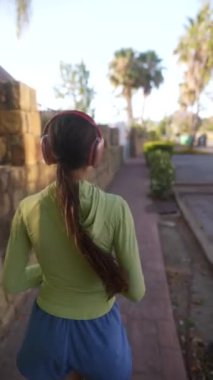 Güneşli bir parkta kulaklıkla koşan, yeşil kapüşonlu ve mavi şortlu genç bir kadının arka görüntüsü..