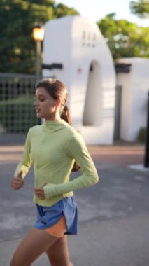 Genç bir kadın güneşli bir günde parkta koşuyor, yeşil bluz ve mavi şort giyiyor, spor ve sağlık için.
