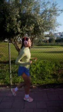 Spor kıyafetli bir kadın güneşli bir günde dışarıda koşuyor, çitlerin ve yeşilliklerin yanında egzersiz yapıyor.