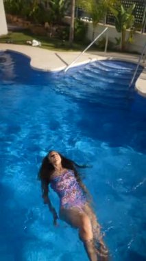 Canlı bir mayo giymiş bir kadın, parlak mavi bir havuzda huzur içinde sırtüstü yüzüyor, güneş ışığını emiyor.