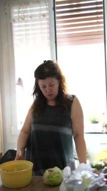 Bir kadın parlak bir mutfakta duruyor, taze sebzeli bir salata hazırlıyor, farklı açılardan görülüyor..
