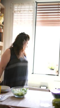 İyi aydınlatılmış bir mutfakta, bir kadın pencerenin kenarında durur, dışarı bakmak için durur, masada kaselerle çevrilidir.