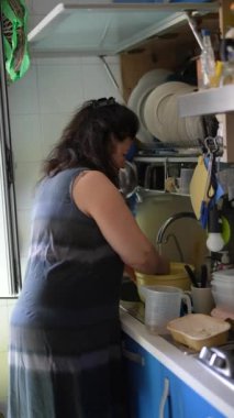 Orta yaşlı bir kadın koyu renk kolsuz bir elbise giyiyor. Mutfakta yemek hazırlayıp bulaşıkları yıkamakla meşgul.