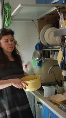 Bir konut mutfağında, bir kadın bulaşık yıkarken ve bulaşık yıkarken çeşitli ev eşyalarını özenle temizler ve düzenler.
