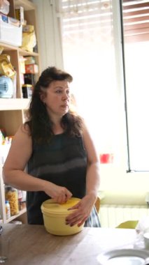 Bir kadın mutfakta, yemek hazırlıyor. Parlak bir pencerenin yakınındaki sarı bir kasede malzemeleri karıştırıyor.