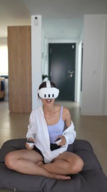 Genç bir kadın sanal gerçeklik deneyiminden zevk alıyor, modern bir evde kontrolörlerle geziniyor..