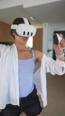Bir kadın evde sanal gerçeklik deneyimine dalıyor, VR kulaklık takıyor ve kontrol cihazlarını tutuyor..
