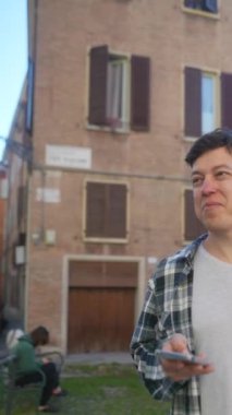 Genç bir adam eski bir şehir binasının yanında duruyor, akıllı telefon kullanıyor, teknolojiye ve iletişime dalmış.