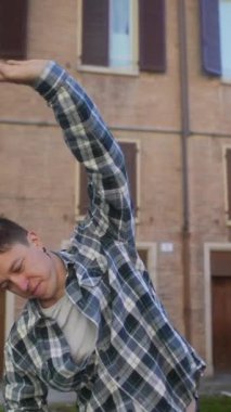 Genç bir adam tuğla binanın dışında, egzersiz rutininin bir parçası olarak ekose gömlek giyerken esniyor.