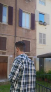 Genç bir adam dışarıda, kahverengi panjurları ve bej duvarları olan eski bir binanın yanında akıllı telefon kullanıyor.