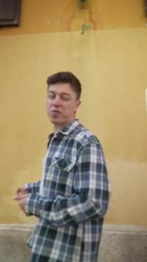 Sıradan bir genç adam, ekose gömlek giymiş, ahşap pencereli ve metal ızgaralı sarı bir binanın yanında yürüyor..