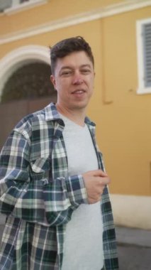 Açık havada turuncu bir binanın yanında, ekose gömlekli ve gri tişörtlü, sıradan bir şehir havasıyla gülümseyen bir adam.