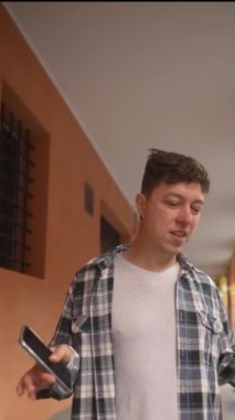 Genç bir adam koridorda yürüyor, sıradan bir kareli gömlek giyiyor ve elinde akıllı telefon tutuyor.