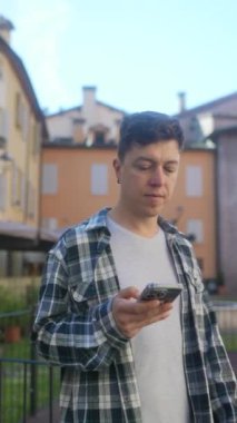 Kareli gömlekli genç bir adam Avrupa 'nın bir kasabasında telefonunu kullanıyor, rahatlamış ve teknolojiye dalmış.