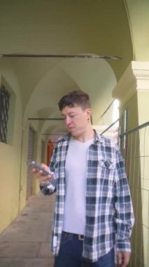 Ekose gömlekli genç bir adam kemerli, örtülü bir yolda yürürken akıllı telefonuna takılır..