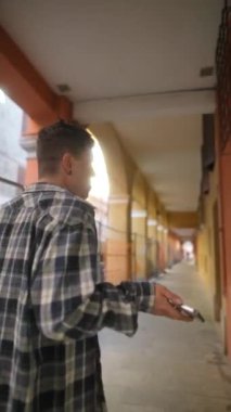 Ekose gömlekli genç bir adamın arka planda canlı bir kemer koridorunda akıllı telefon kullanarak yürürken görüntüsü..