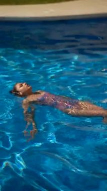 Genç bir kadın yüzme havuzunda sırt üstü yüzüyor güneşli bir günün tadını çıkarıyor ve suda dinleniyor..