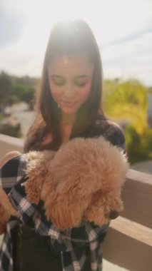 Genç bir kadın gün ışığında güneşlenirken sevimli tüylü köpeğini balkonda şefkatle tutuyor.