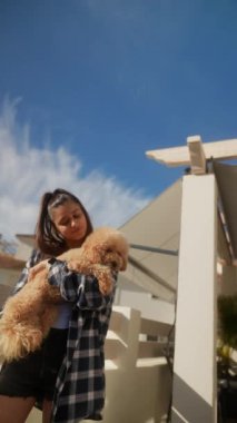 Genç bir kadın neşeli bir şekilde tüylü bir köpek tutuyor, ikisi de güneşli bir günde açık mavi bir gökyüzünün altında mutlu anları paylaşıyor.