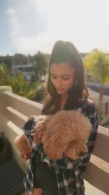 Genç bir kadın balkonda pofuduk bir köpek tutuyor ve gülümseme ve rahatlıkla güneşli bir günün tadını çıkarıyor.