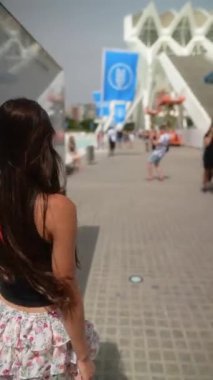 Genç bir kadın güneşli bir günde çağdaş bir şehir bölgesinde yürüyor, modern mimari ve kültürü yakalıyor..