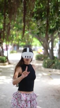 Genç bir kadın parkta, beyaz bir VR kulaklıkla artırılmış gerçekliği yaşıyor, gülümseme takıyor.