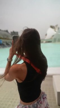 Uzun saçlı ve güneş gözlüklü bir kadın, modern bir açık hava sahasında akıllı telefonunu kullanarak fotoğraf çekiyor..