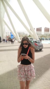 Modern bir şehir bölgesinde güneş gözlüklü genç bir kadın özel mimarisi olan akıllı bir telefon kullanıyor..