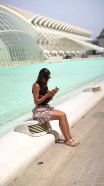 Güneşli bir günde fütüristik mimari tasarımı olan bir kadın akıllı telefonunu modern su özelliklerinin yanında kullanıyor.