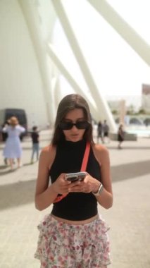 Yaz kıyafetleri giymiş genç bir kadın, modern mimarinin arka planına sahip açık havada akıllı bir telefon kullanıyor.