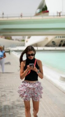 Siyah bluzlu ve çiçekli etekli bir kadın modern mimari ve su özellikleriyle yürür ve akıllı telefon kullanır..