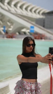 Genç bir kadın modern mimari bir binanın yakınındaki havuz kenarında selfie çekiyor, güneşli bir günün tadını çıkarıyor.