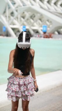 Şık bir genç kadın, havuz kenarında boş zamanlarının keyfini çıkarırken VR kulaklık takıyor ve kumanda tutuyor.