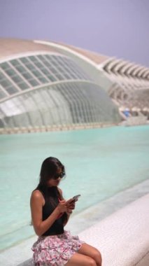 Çiçekli etek giyen uzun saçlı genç bir kadın, akıllı telefonunu fütüristik mimari ve su ile kullanır..