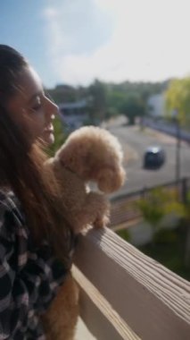 Genç bir kadın pofuduk köpeğini tutarken güneşli bir balkonda manzaralı bir manzaranın tadını çıkarıyor..