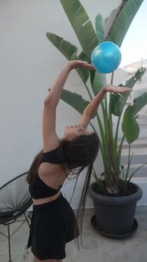 Genç bir kadın zarif bir şekilde dışarıda mavi bir topla ritmik jimnastik yapıyor, yeteneklerini ve zarafetini sergiliyor.