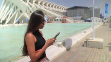 Genç bir kadın, modern mimari ortamda güneşli bir günde çağdaş bir havuz tarafından akıllı telefonuna sarılmış.