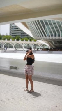 Gündelik yaz kıyafetleri giymiş genç bir kadın modern binaların ortasında güneşli bir günün tadını çıkarırken mutlu görünüyor.