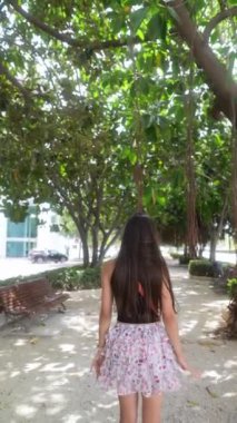 Uzun saçlı bir kadın ağaçlarla çevrili güneşli bir parkta yürüyor ve huzurlu bir ortam yaratıyor. Arkadan görüntüle