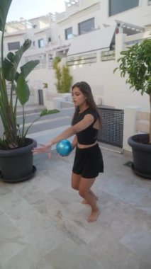 Genç kız dışarıda mavi topla oynuyor, çeşitli atletik hareketler yapıyor ve dışarıda eğleniyor..