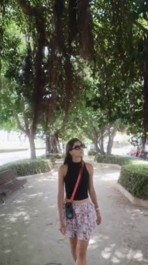Şık bir kadın ağaçların altında yürüyor, güneş gözlüğü takıyor ve parkta rahat bir yürüyüş için şık bir yaz kıyafeti giyiyor.