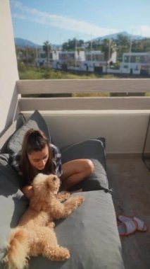 Bir kadın balkonunda güneşli bir günün tadını çıkarır, rahat bir kanepede tüylü köpeğiyle oynar ve dinlenir..