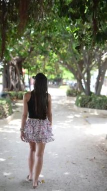 Bir kadın güzel yeşil bir parkta geziyor, çiçekli bir elbise giyiyor ve doğada bir yaz gününün tadını çıkarıyor.