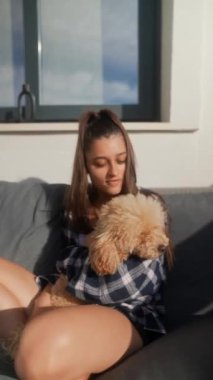 Genç bir kadın sevgisini, tüylü köpeğini sıcak güneş ışığı altında açık bir kanepede sevgiyle kucaklayarak gösterir.