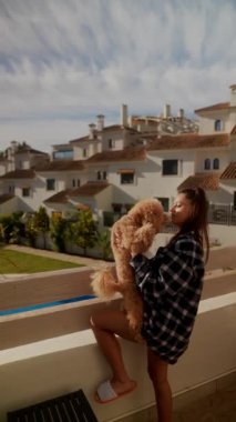 Bir kadın balkonda tüylü bir köpeği sevgiyle kucaklıyor, açık güneşli bir gökyüzünün altındaki evlerin manzarasının tadını çıkarıyor.