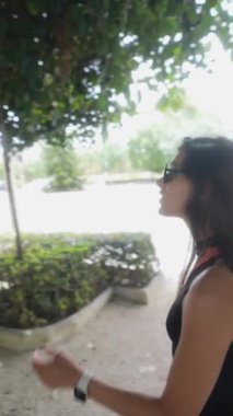 Güneş gözlüklü ve uzun saçlı bir kadın parkta güneşli günün ve canlı yeşilliğin tadını çıkarıyor.