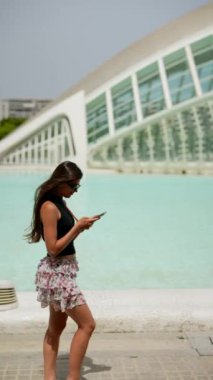 Genç bir kadın modern binaların ve su hatlarının yanında yürüyor, mimari bir ortamda telefonuna takılıp kalıyor.